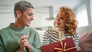 mother and adult daughter sitting on a couch; the daughter holds a gift she has just received
